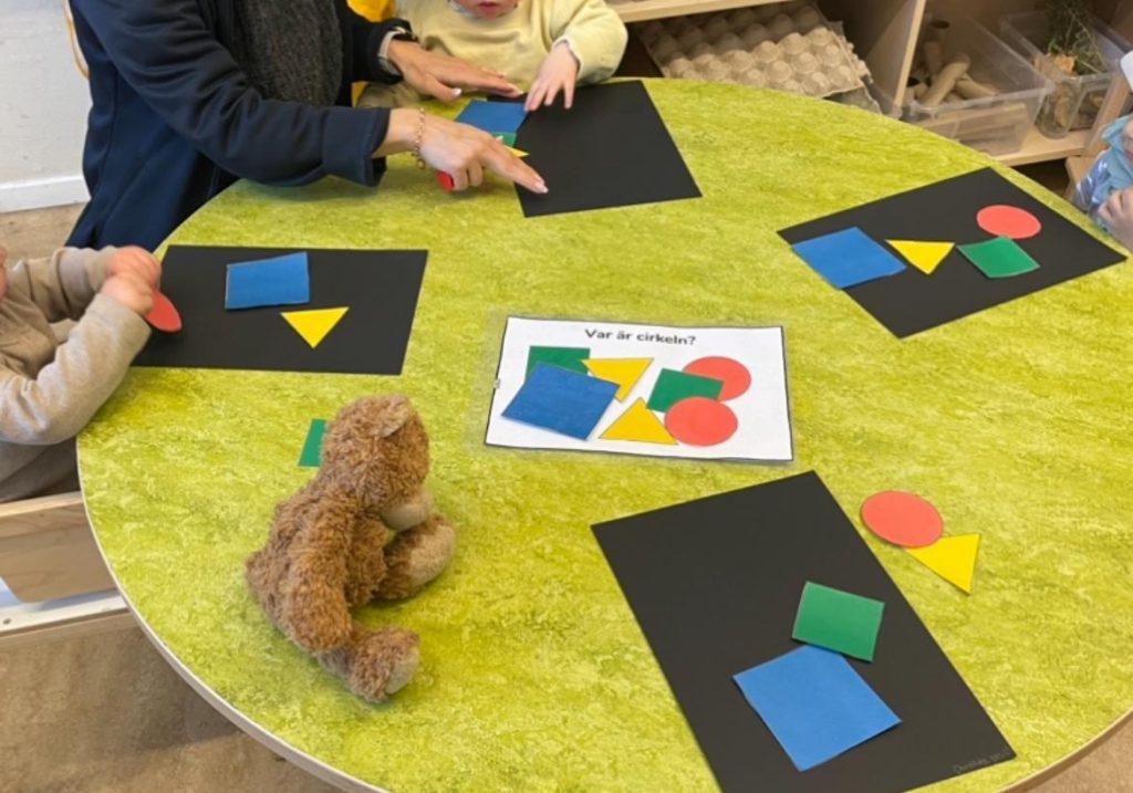 Ett barn och en vuxen sitter vid ett runt bord täckt med grön filt, med svarta papper där geometriska former i olika färger ligger utplacerade, och en brun nallebjörn i mitten