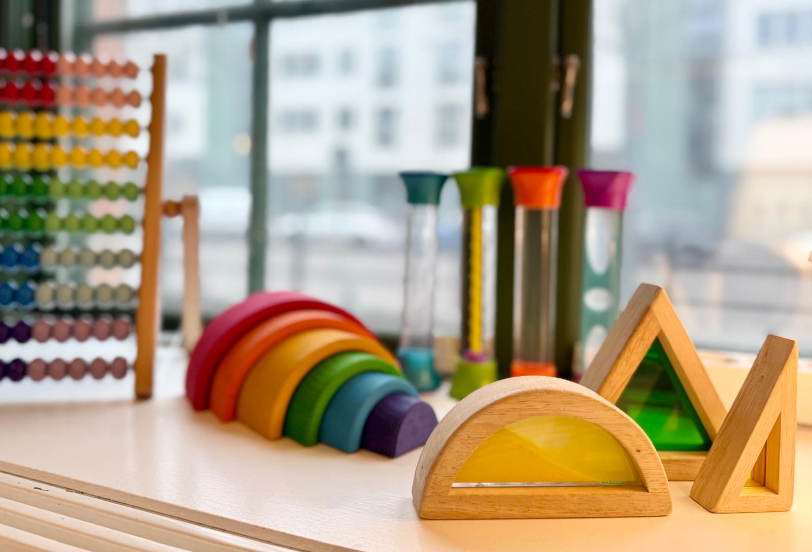 Pedagogiska träleksaker uppställda på en fönsterbräda: en kulram i regnbågsfärger, en trärabåge av staplade halvcirklar, färgade timglas samt geometriska träformer med färgat glas. Suddig stadsvy syns genom fönstret i bakgrunden.