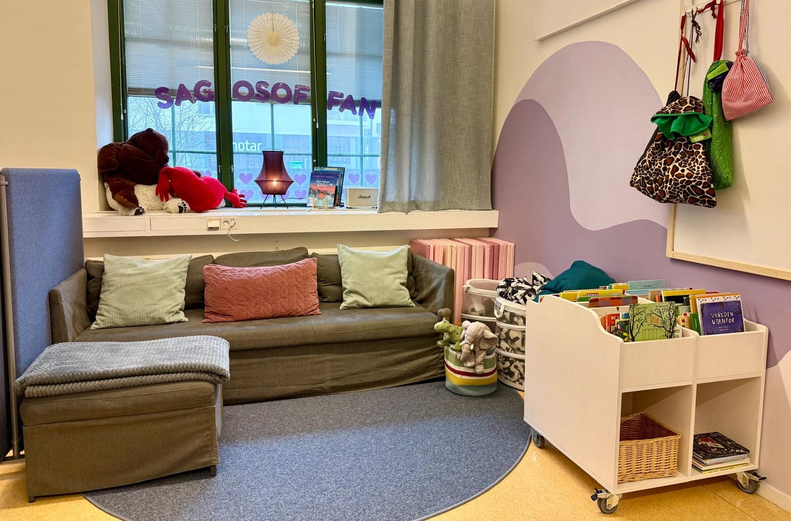 Mysig läshörna på förskola med grön soffa, kuddar och en rund matta. På väggen bakom finns en lila bergsmålning och krokar med väskor, medan en rullbar bokhylla med barnböcker står bredvid. I fönstret hänger bokstäver som bildar ordet 'Sagosoffan.