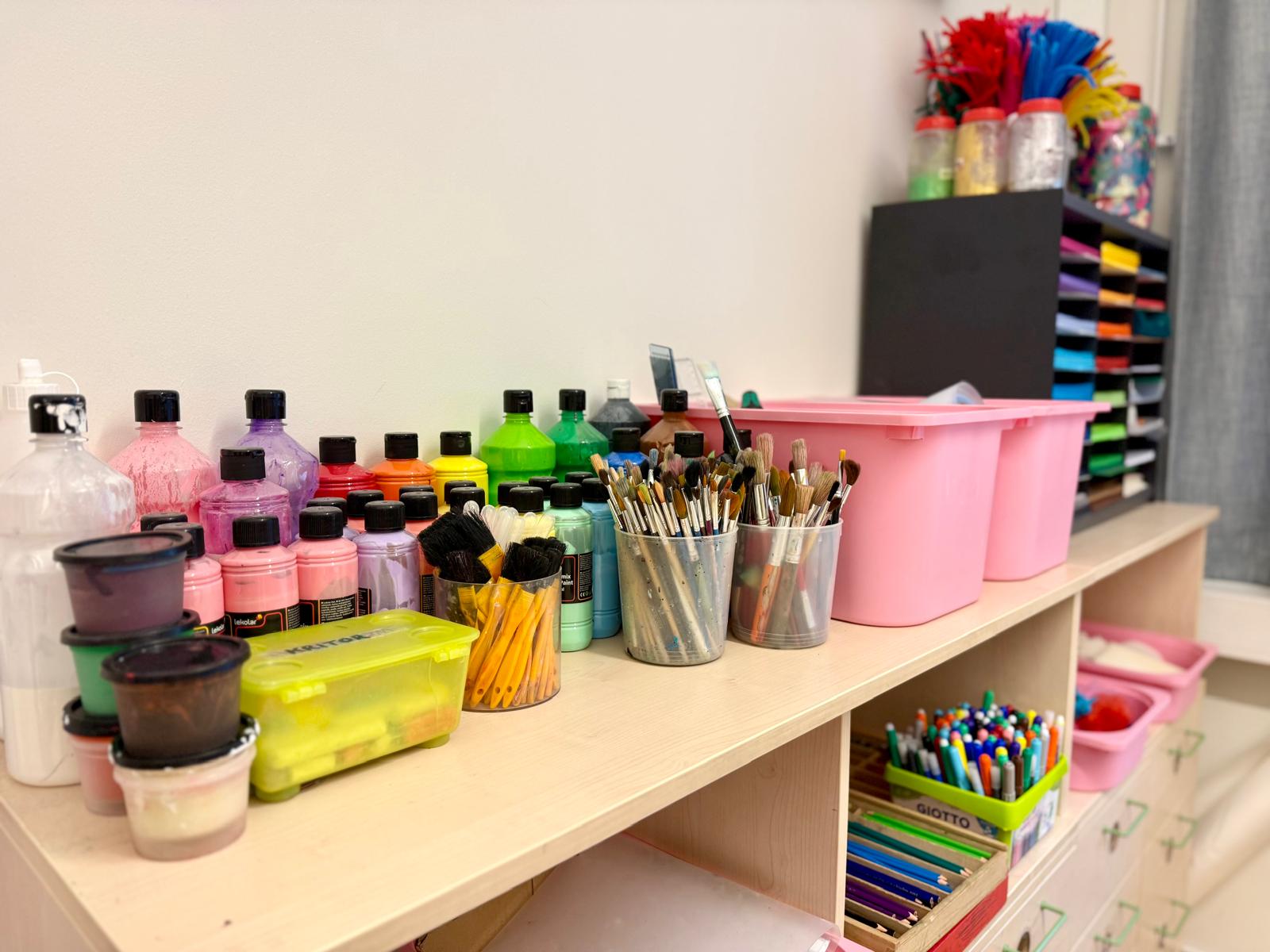 Hylla i en förskolas skaparrum fylld med färgglada materialflaskor, penslar i olika burkar, pennor, kritor och rosa förvaringslådor. Underliggande fack innehåller pennor och kritor i förvaringsboxar.