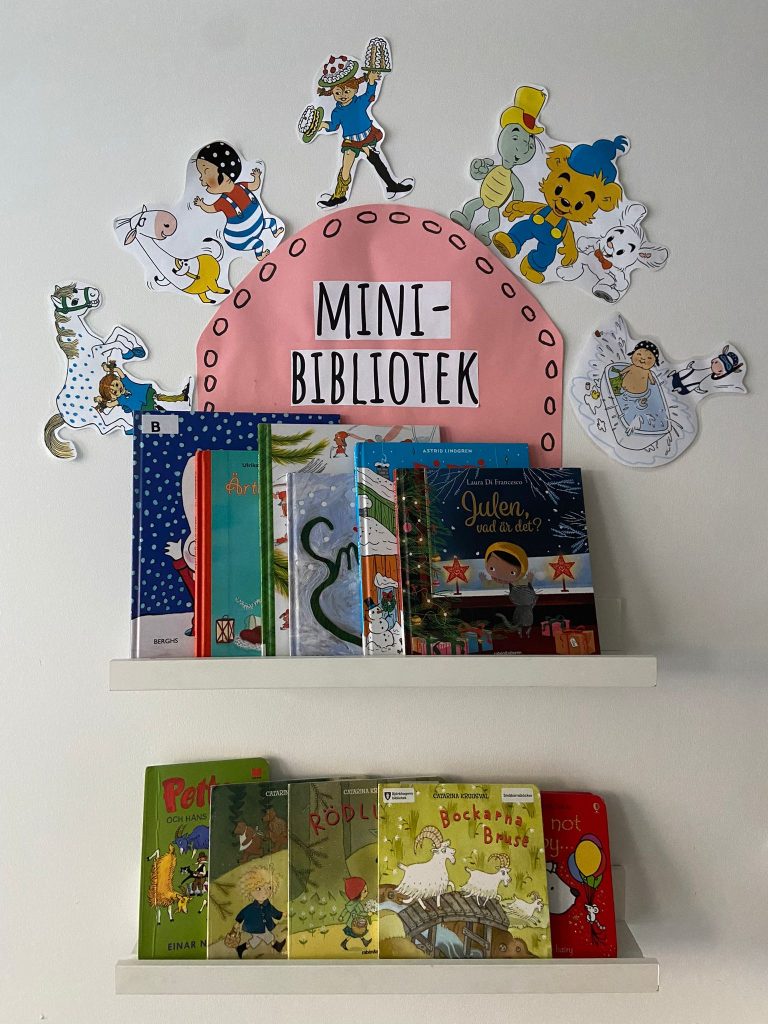 n vägganordning med texten "Mini-bibliotek" på rosa bakgrund, dekorerad med figurer från svenska barnböcker, ovanför två hyllor med barnböcker uppställda på förskolan.