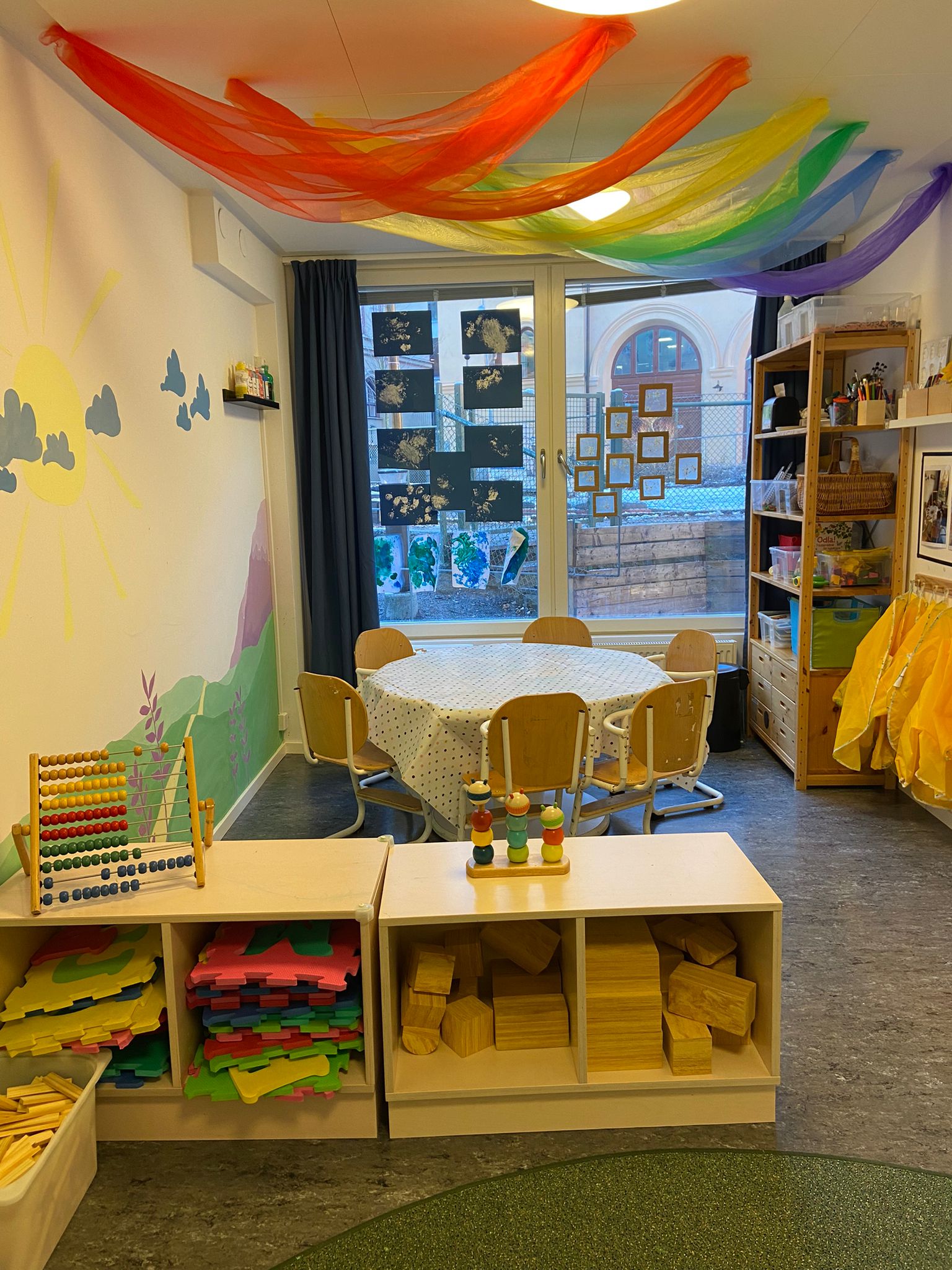 Förskolerum med regnbågsfärgade tygbanor draperade i taket, ett runt bord med stolar täckt av en prickig duk, och hyllor fyllda med byggklossar, pussel och leksaker. På väggen finns en målning av sol och moln, och ett fönster med barnens konstverk syns i bakgrunden