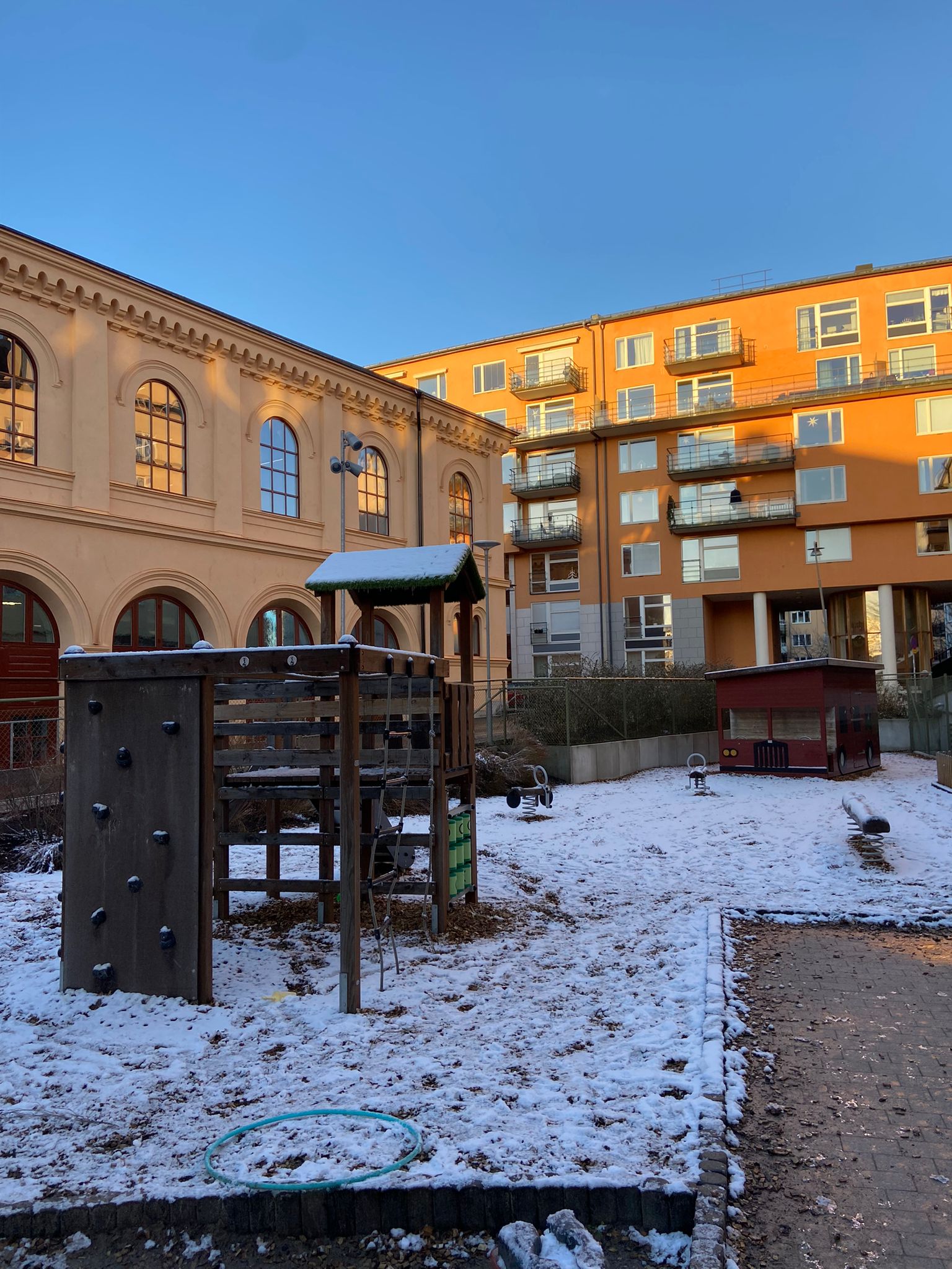 Snötäckt lekplats med klätterställning i trä, gungdjur på fjädrar och en liten röd lekstuga formad som en bil. I bakgrunden syns en gul tegelbyggnad med valvfönster samt ett orange flerfamiljshus under klarblå himmel.