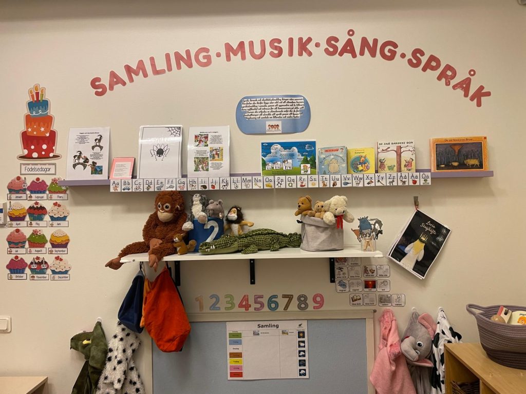 Samling, Musik, Sång, Språk' i rött, dekorerad med alfabetsplanscher, barnböcker och en hylla med gosedjur som apa, krokodil och björnar. Nedanför finns siffror, en samlingstavla och en väggdekoration för barnens födelsedagar.