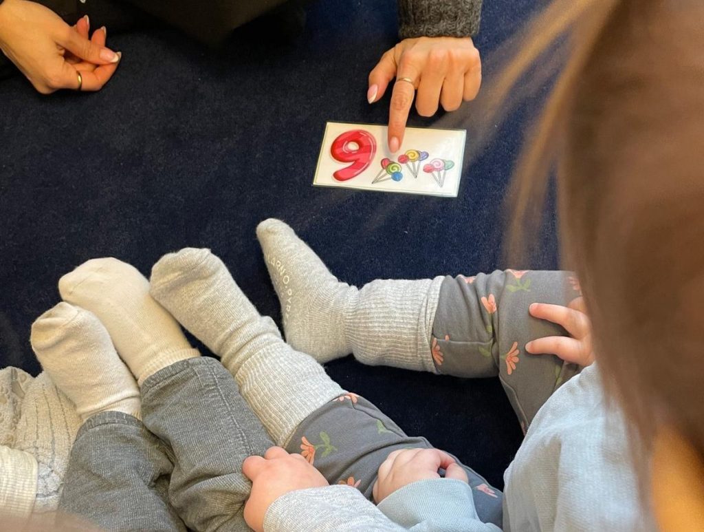 Vy uppifrån ett antal barn som sitter i ring på en mörkblå matta, där en vuxen pekar på ett kort med siffran 9 och tillhörande illustrationer. Barnens ben och strumpfötter syns i förgrunden.